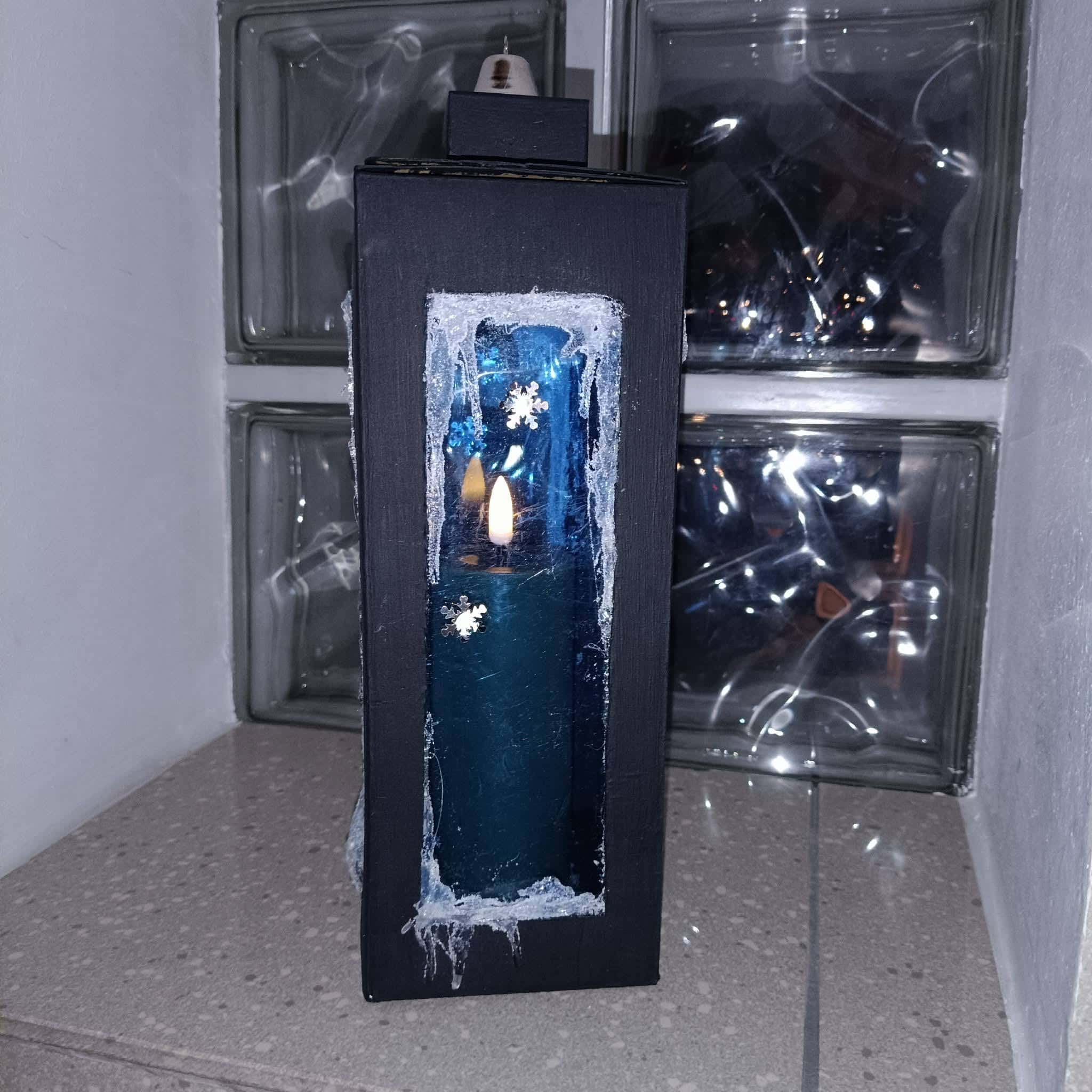 Czarny lampion z wyciętym okienkiem, niebieską świecą i zimową dekoracją ze śnieżynkami – praca dziecka.