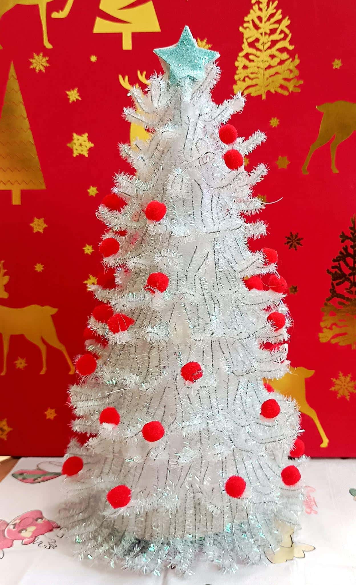 Biała choinka z drucików kreatywnych ozdobiona czerwonymi pomponami i gwiazdką na czubku – praca dziecka.