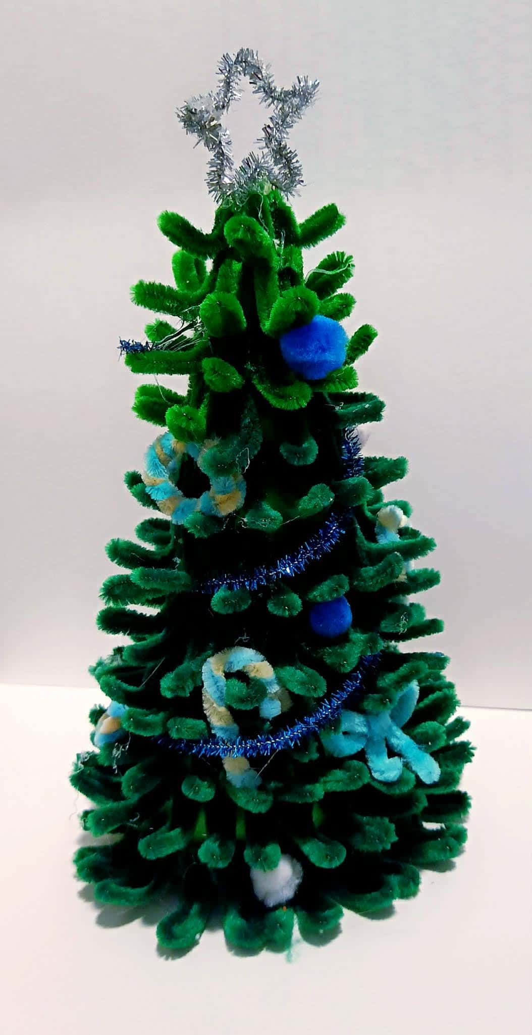 Przestrzenna choinka wykonana z zielonych drucików kreatywnych, ozdobiona niebieskimi i jasnymi pomponami oraz srebrną gwiazd